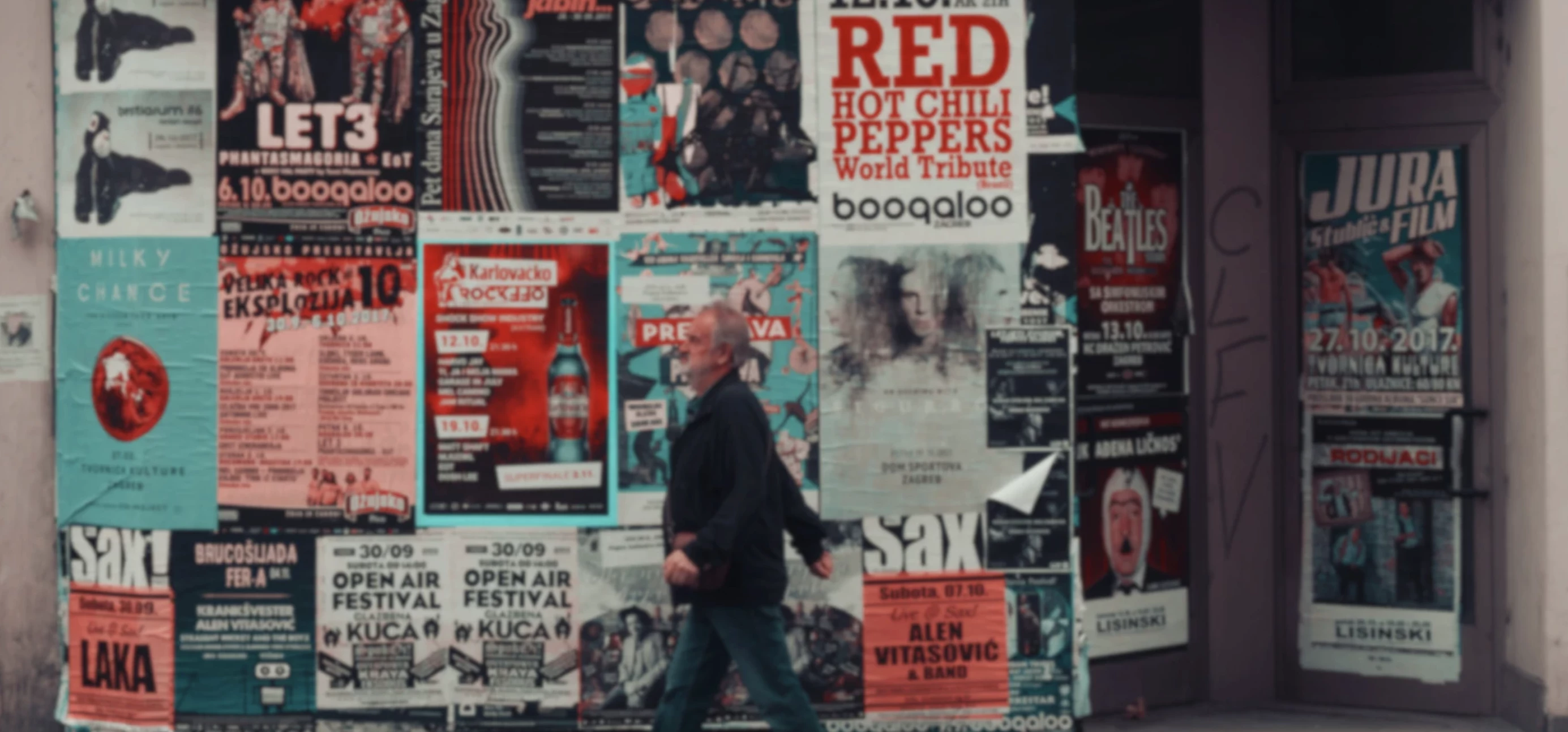 Man with grey hair and beard walks left in front of a wall of music posters